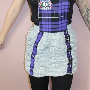 May include: A purple and black plaid overall skirt with a skull and flower patch. The skirt has a ruffled bottom and is paired with a black shirt.