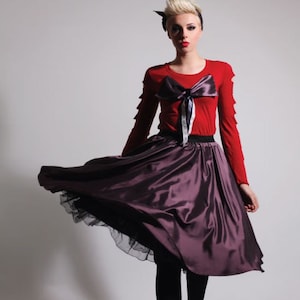May include: A woman wearing a red long-sleeved shirt with a large bow on the front and a purple satin skirt. The skirt has a black tulle underskirt.