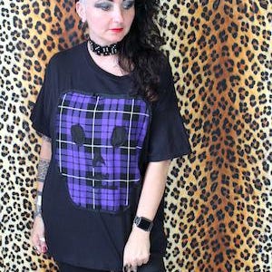 May include: Black t-shirt featuring a purple and black plaid skull design. The shirt has short sleeves and a loose fit. The background is a leopard print pattern.
