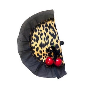 Op de afbeelding: Een halfrond stoffen accessoire met een opvallend luipaardpatroon in geel en zwart. De rand is afgewerkt met een brede, geplooide zwarte tule ruche. Een kleine zwarte fluwelen strik en twee glanzende rode kersen zijn bevestigd.