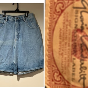 May include: Light blue denim shorts hanging on a black hanger. The shorts have a high waist, front pockets, and a button closure. The shorts are made of a light wash denim material. A label with text is visible on the right.