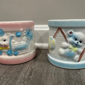 Puede incluir: Dos macetas de cerámica con temática de bebé, una rosa con un cochecito azul y un conejito blanco, la otra azul con un osito de peluche blanco.