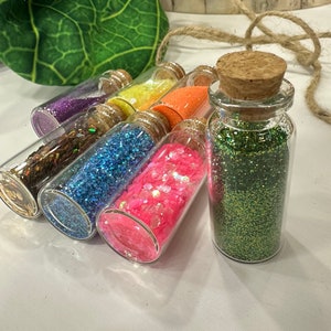 Puede incluir: Siete pequeñas botellas de vidrio con tapas de corcho llenas de purpurina de diferentes colores. Las botellas están dispuestas en una fila sobre una superficie blanca.