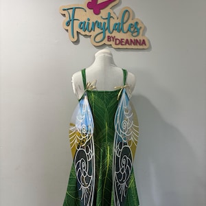 May include: A green fairy dress with leaf patterns and attached iridescent wings. The wings have a white, yellow, and blue design. The dress is displayed on a mannequin. The sign above the dress reads "Fairytales by Deanna".