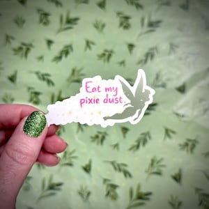 May include: A white sticker with the text "Eat my pixie dust" in pink, featuring a silhouette of a fairy. The sticker is held against a light green background with a leaf pattern.