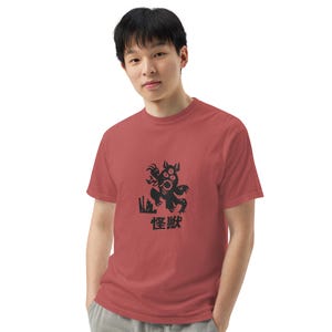 Puede incluir: Una camiseta roja con un gráfico negro de un monstruo de tres ojos con el texto "怪獣" en japonés.