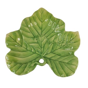 May include: A green ceramic serving dish shaped like a leaf with a vine design. The dish has a hole in the center.