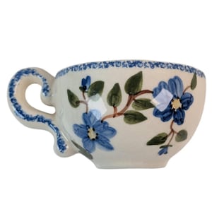 May include: A ceramic teacup with a white base, adorned with hand-painted blue floral designs and green leaves. The rim and handle are detailed with blue accents. The cup has a curved handle and a rounded shape, perfect for enjoying tea or coffee.