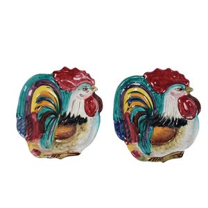 以下が含まれることがあります： 鶏の形をしたセラミック製のサービングディッシュ2つ。ディッシュは、赤、黄、緑、青、黒など、明るい色で描かれています。鶏は白い体で、赤い鶏冠と肉垂があり、尾は扇形に広がっています。