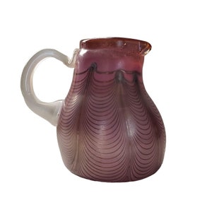 Op de afbeelding: Een kleine, paarse glazen kan met een heldere glazen handvat. De kan heeft een draaiende patroon van dunne lijnen.