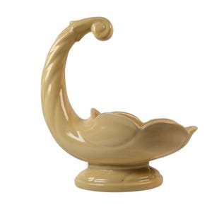 May include: A pale yellow ceramic bowl featuring a high, curved, twisted handle that ends in a spiral scroll. The bowl has a scalloped, petal-like rim and rests on a circular pedestal base, presenting a smooth, glossy finish throughout the piece.
