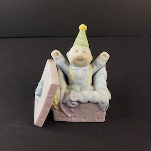 May include: A porcelain figurine of a Cabbage Patch Kid wearing a blue and yellow outfit and a green and yellow party hat. The figurine is sitting in a pink gift box with a blue ribbon.