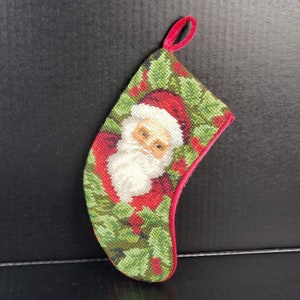 Könnte beinhalten: Ein roter und grüner Weihnachtssocken mit einer Nadelbild-Darstellung von Santa Claus' Gesicht, umgeben von Stechpalmenblättern und Beeren.
