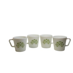 May include: Four white ceramic mugs with a green floral design. Each mug has a handle and a slightly flared shape. The floral pattern is a repeating design of small flowers and leaves. The mugs are arranged in a row.