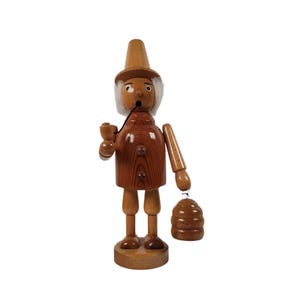 May include: A wooden figurine of a person with a white beard and hat, holding a pipe and a small wooden basket. The figurine is painted in shades of brown and has a rustic appearance.