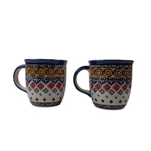 May include: Two ceramic mugs with a blue rim and handle. The mugs feature a repeating pattern of orange, blue, and red geometric shapes. The mugs are white with a dark blue rim and handle.
