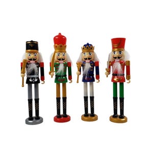 Puede incluir: Cuatro cascanueces de madera en varios colores. Cada figura tiene un uniforme diferente, incluyendo negro, verde, azul y rojo. Tienen pelo y bigote blancos, y están sobre una superficie blanca.