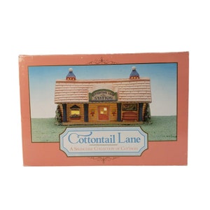Puede incluir: Una casita en miniatura decorativa con el texto "Cottontail Lane Station" en el tejado. La casita está en una caja rosa con el texto "A Springtime Collection of Cottages".