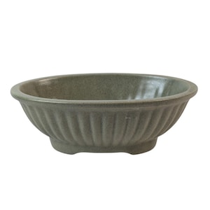 May include: A sage green ceramic bowl with a fluted exterior. The bowl has a rounded oval shape and sits on three small feet. The rim is smooth, and the bowl's surface has a speckled texture.