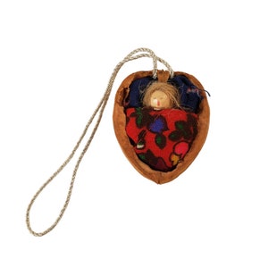 May include: A small doll made of wood and fabric is sleeping inside a heart-shaped walnut shell. The doll has blonde hair, a red and blue patterned blanket, and a string attached to the shell.