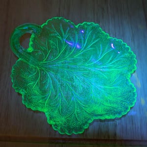 May include: A glowing green glass leaf-shaped dish with a handle. The dish has a detailed leaf vein pattern and a scalloped edge. The glass appears to be vintage and glows brightly under a black light.