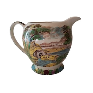 May include: A white ceramic jug with a handle and a scenic design of a watermill, trees, and mountains in shades of green, brown, and yellow.