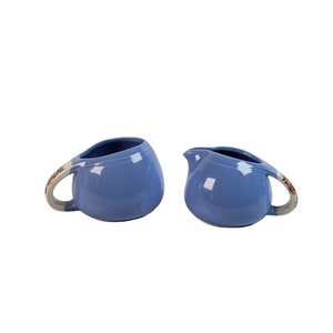 May include: A set of two blue ceramic creamer and sugar bowls with floral accents. The bowls are round and have a slight curve to them. The handles are white with floral designs.