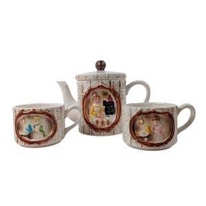 Può includere: Un servizio da tè vintage con una teiera e due tazze. Il set in ceramica color crema ha un motivo strutturato e scene in rilievo incorniciate in marrone di donne. La teiera ha un pomello e un beccuccio marrone. Le tazze hanno manici.