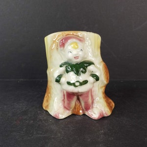 May include: A ceramic planter shaped like a tree stump with a small elf figurine standing in front of it. The elf is wearing a green hat, a green and white jacket, a brown belt, and pink pants. The planter is painted in shades of brown, green, and pink.