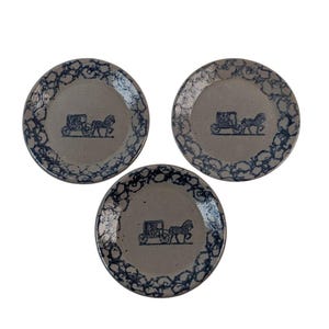 May include: Three round ceramic plates with a light tan centre and a dark blue sponge-painted rim. Each plate features a dark blue graphic in the centre depicting a horse-drawn carriage with a single horse pulling a four-wheeled passenger vehicle.