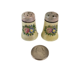 Puede incluir: Un par de saleros y pimenteros vintage con un diseño floral. Los saleros son blancos con flores rosas y azules y hojas verdes. Los saleros son aproximadamente del tamaño de una moneda de 20 céntimos de euro.