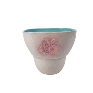 May include: A ceramic vase with a light beige exterior and a turquoise interior. The vase features a textured surface and a pink floral design on the front. The vase has a unique shape, with a wider top and a narrower base.