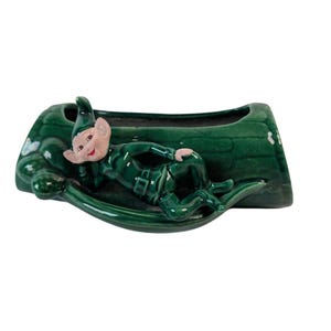 May include: A green ceramic planter featuring a reclining elf figure. The elf is dressed in a green outfit with a pointed hat and rosy cheeks. The planter has a boat-like shape with a textured exterior.