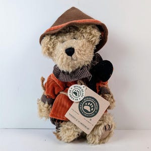 May include: A plush teddy bear with curly beige fur wearing a brown hat with an orange trim, a rust-colored corduroy jacket, and a brown and white checkered collar. A beige tag attached to the bear reads Boyds Bears and Einstein Q. Scaredybear.