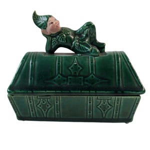 May include: A green ceramic trinket box with a reclining elf figurine on top. The box is rectangular with a hinged lid and decorative diamond and rivet details. The elf has a pink face and is wearing a green leaf-themed outfit.