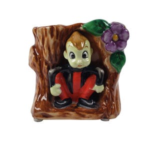 Peut inclure: Pot de fleurs vintage en céramique en forme de tronc d'arbre, avec une figurine de dessin animé assise en vêtements rouges et noirs. Une fleur violette et des feuilles vertes ornent le côté. Le pot est marron avec une finition brillante.