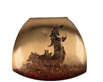 May include: A gold-toned, shield-shaped compact mirror. The mirror features an engraved design of a Viking ship with a figurehead and floral accents. The compact has a slightly curved shape and a smooth surface.