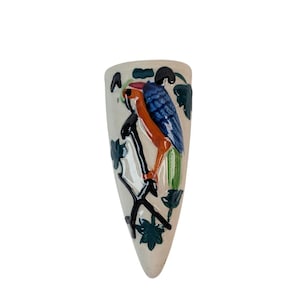 May include: A cream-colored ceramic wall pocket vase with a painted parrot design. The parrot has blue wings, orange body, and green tail, perched on a black branch with green leaves. The vase is shaped like a long, narrow teardrop.
