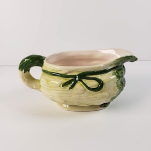 Puede incluir: Una jarra de crema de cerámica con forma de col, con un asa verde y una cinta verde atada alrededor de la parte superior. La jarra está pintada en tonos verdes y amarillos.