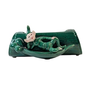 May include: A vintage ceramic planter in the shape of a green log with a reclining elf figure. The elf is wearing a green outfit with a white face and is resting on a green leaf-shaped base. The planter has a glossy finish.