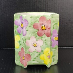 May include: A green ceramic planter with a hexagonal shape and a floral design. The planter features pink, purple, and yellow flowers with green leaves.