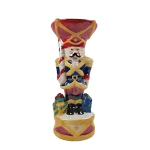 May include: A ceramic nutcracker candle holder with a red hat, blue and gold uniform, and a candy cane in his hand. He is standing on a gold base with a star.