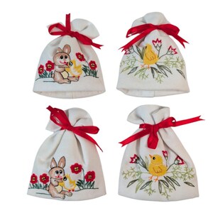 May include: Four cream-colored fabric bags tied with red ribbons. Each bag features embroidered designs: two with a bunny and chick, and two with a chick. The bags are decorated with red flowers and green foliage, perfect for Easter.