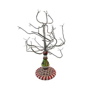 May include: A decorative wire tree with a whimsical design. The tree features a silver-colored wire structure with a detailed base. The base has a red and white striped pattern with a green and gold decorative element. The tree is designed for displaying small ornaments.