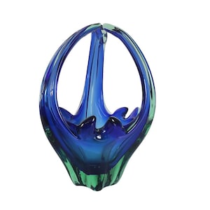 Puede incluir: Una cesta de vidrio soplado a mano en tonos azules y verdes. La cesta presenta un asa y una forma orgánica y acampanada. La base es de un verde vibrante, que transiciona a un azul profundo en el centro y a un azul más claro en la parte superior.