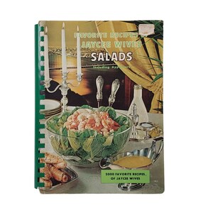 May include: A vintage cookbook with a green spine titled "Favorite Recipes of Jaycee Wives Salads Including Appetizers" with a photo of a salad bowl with shrimp and lettuce on a table with candles and silverware. The bottom of the cover reads "2000 Favorite Recipes of Jaycee Wives".