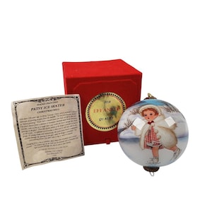 Puede incluir: Bola de Navidad Patsy Ice Skater pintada a mano con caja roja y etiqueta. El adorno de cristal presenta a una niña patinando sobre hielo en una escena invernal. La caja tiene un sello dorado que dice "For Effanbee Quality".