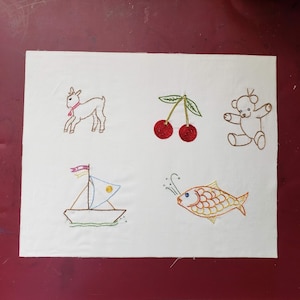 Peut inclure: Un tissu blanc avec des images brodées d'une chèvre, de deux cerises, d'un ours en peluche, d'un voilier et d'un poisson. Les images sont réalisées dans différentes couleurs, notamment le rouge, le jaune, le bleu et le vert.