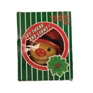 Puede incluir: Una decoración navideña iluminada de color rojo y amarillo con forma de cara sonriente que lleva un sombrero rojo. La caja dice "Let There Be Light!" y "Battery Operated Wall Hanging Decoration Lamp".
