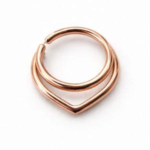 Op de afbeelding: Een roségouden septumring met een dubbellaags ontwerp. De bovenste laag is een complete cirkel, terwijl de onderste laag een V-vorm vormt. De sieraden zijn geplaatst tegen een witte achtergrond.